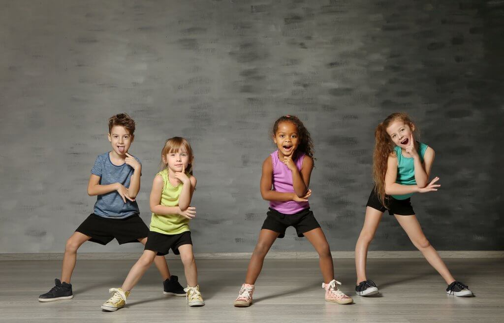 Group of multi cultural kids pose towards the camera with their hands pointing upwards on their faces in a pondering pose. Their legs are spaces out in a bent stance. Grey wall and wooden floor surrounds them.