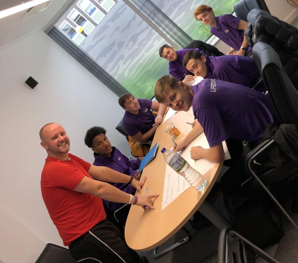 Group of men in CVLife purple clothing and red shirt sit around a table, looking towards the camera. They are mid learning for their Modern Apprenticeship.