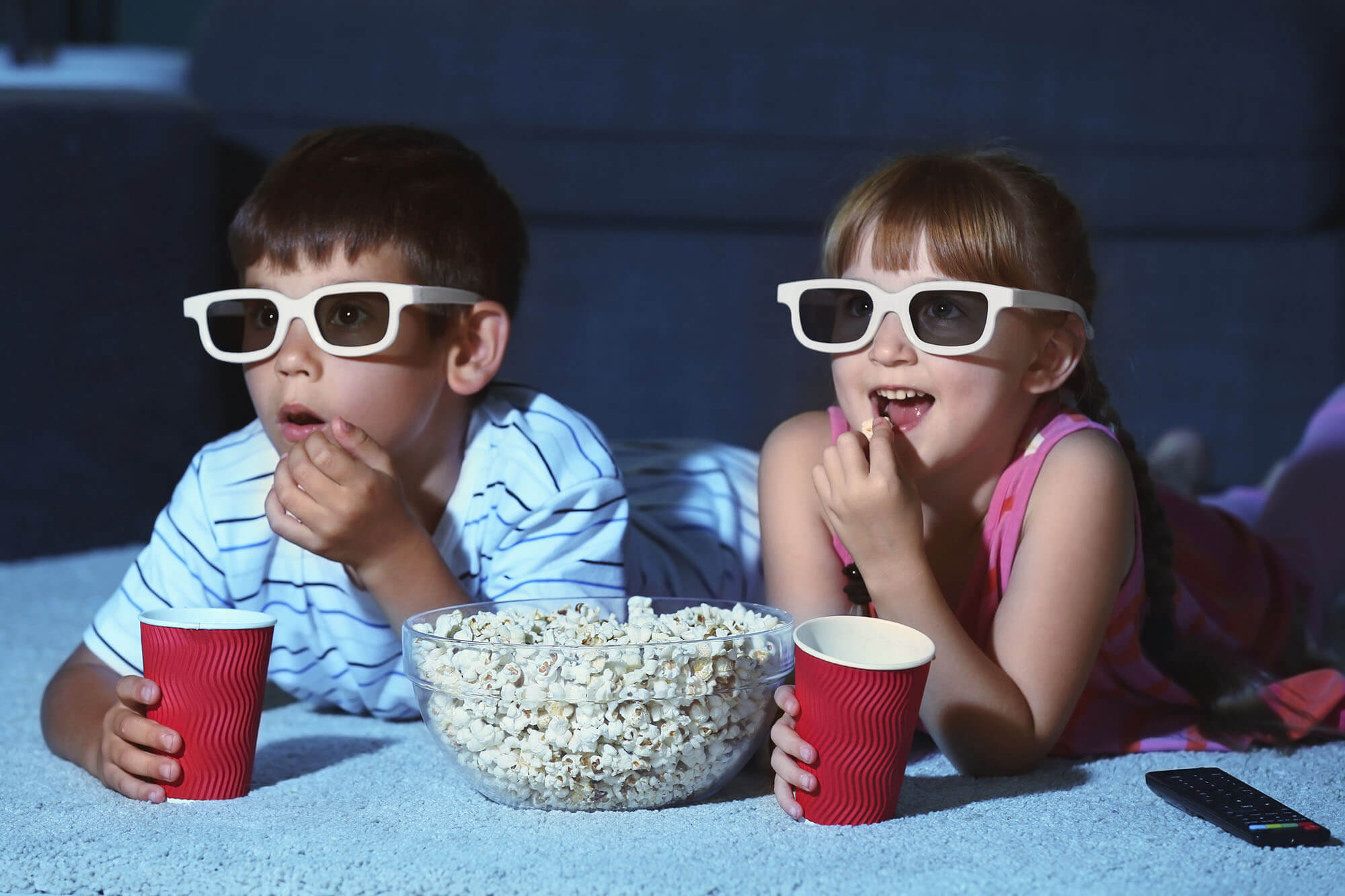 Boy and girl lay down with white 3D glasses over their face. A bowl of popcorn is in front of them along with a remote. The both hold red cups in their hands.
