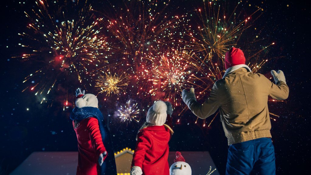 A man, woman and child in coasy clothing stand outside, looking towards fireworks that light up the sky