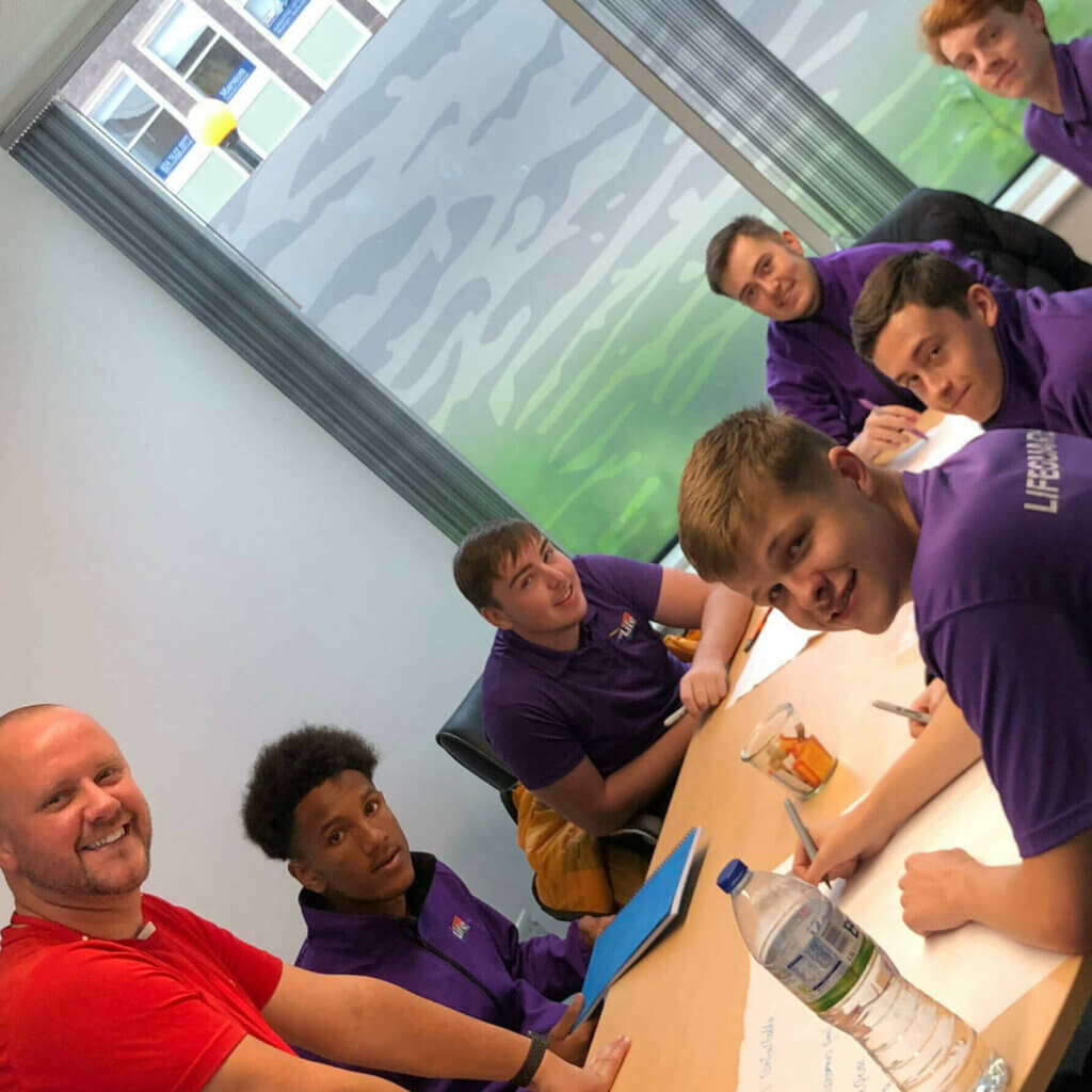 Group of men in CVLife purple clothing and red shirt sit around a table, looking towards the camera. They are mid learning for their Modern Apprenticeship.